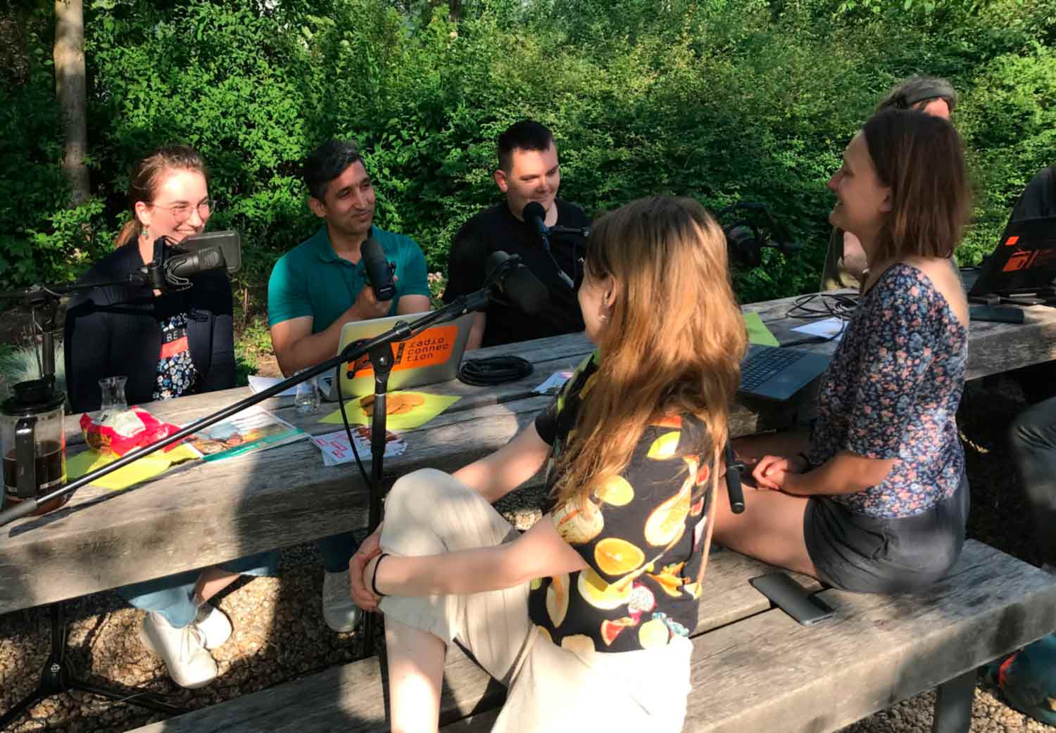 drei Frauen und zwei Männer sitzen um einen Holztisch, draußen im Grünen und haben Mikrofone vor sich.
