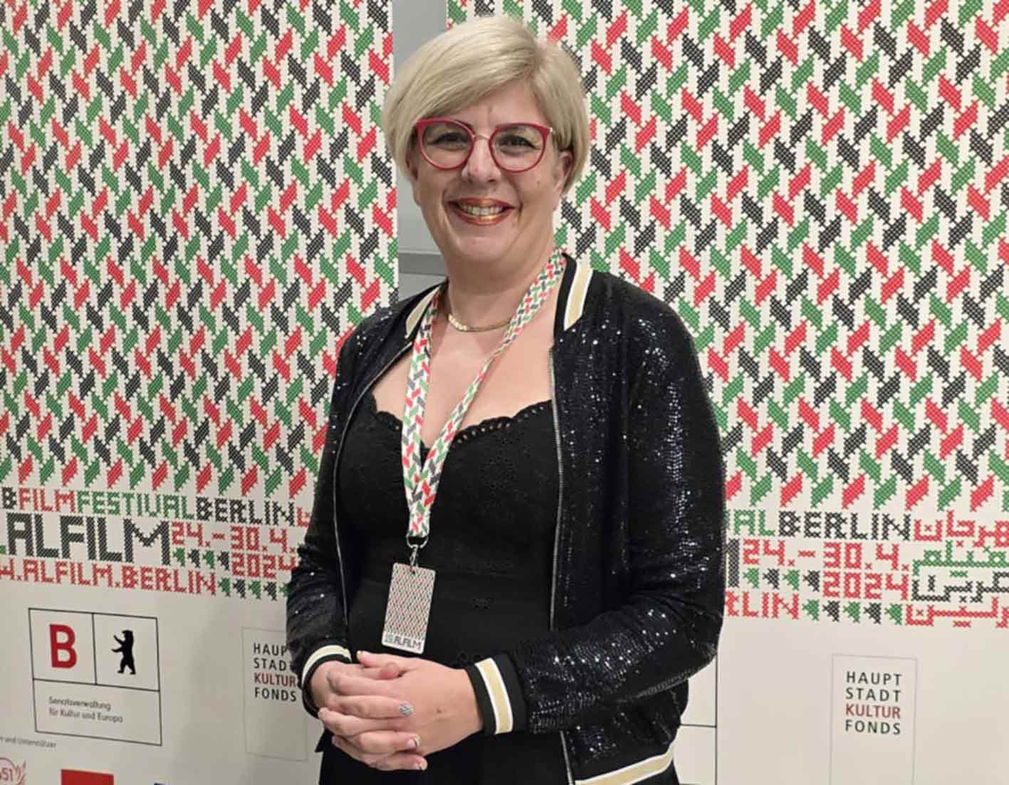 Eine festlich gekleidete Frau steht vor den Plakaten des Filmfestivals ALFILM auf denen ein Muster in rot, grün schwarz und weiß zu sehen ist.
