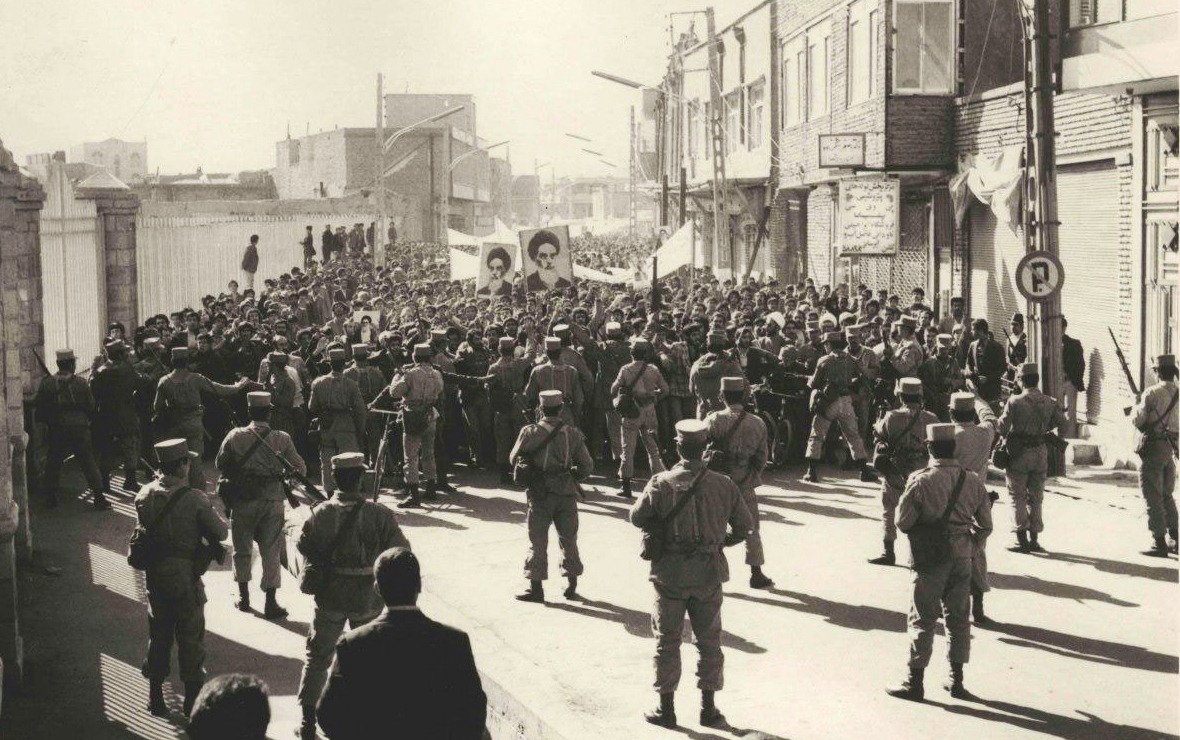 Im Vordergrund stehen Soldaten auf die eine große Demonstration zukommt. Die Demonstranten tragen Fotos von Chomeini.