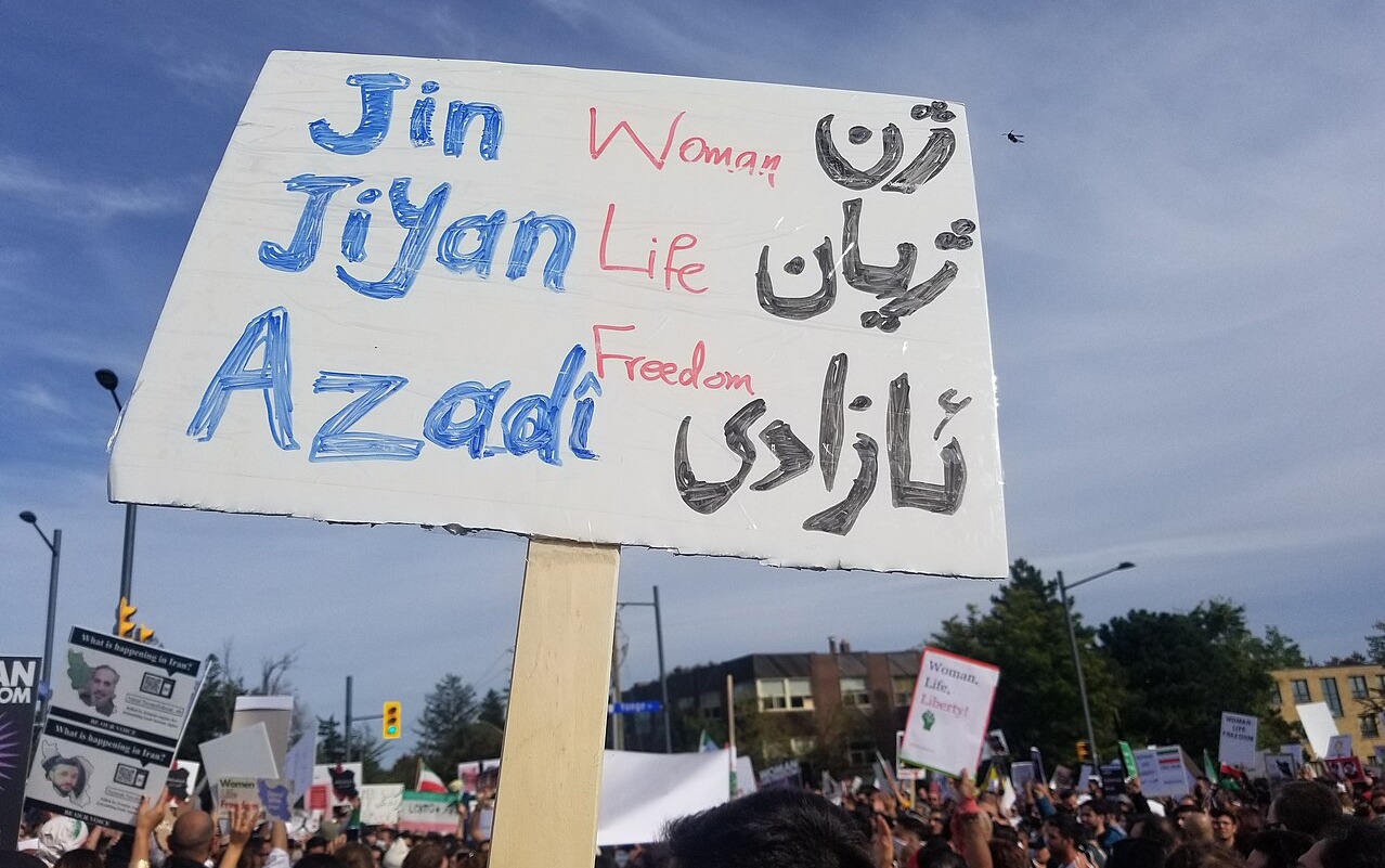 Eine Demonstration, im Vordergrund ein Schild mit: Woman, Life, Freedom (Jin, Jiyan, Azadi) auch kurdisch und englisch