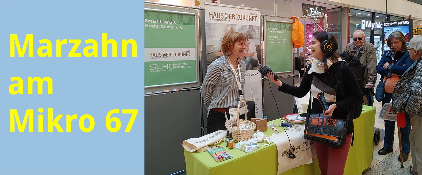 Hinter einem Tisch mit Flyern, T-Shirts und Luftballons steht eine Frau und spricht in ein Mikrofon, das ihr eine Frau mit Kopfhörer, die vor dem Tisch steht, entgegenstreckt. Im Hintergrund hängt, an einer Stellwand, ein Plakat mit der Aufschrift Haus der Zukunft.