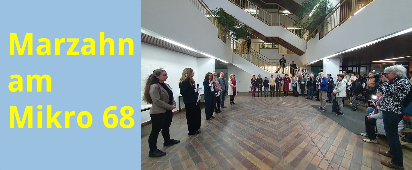 In einem großen Foyer mit Treppenhaus stehen sechs Leute in einer Reihe und haben jeweils ein Mikrofon in der Hand. Gegenüber steht eine große Gruppe von Menschen, der mehrstöckige Raum in der dazwischen ist frei.