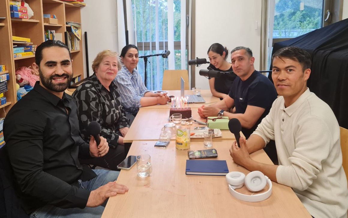 drei Frauen und drei Männer sitzen an einem Tisch. Sieben jeweils ein Mikrofon vor sich. Auf dem Tisch steht ein Schokoladenkuchen mit brennenden Kerzen, daneben liegt ein eingepacktes Geschenk mit einer Schleife drumherum.